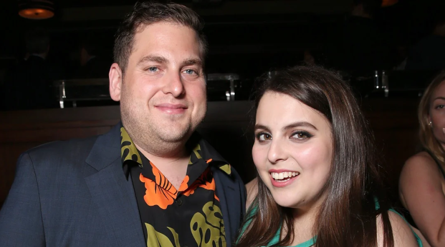 A man in a dark suit and colorful shirt smiles next to a woman in a green dress, both posing for a photo at an indoor event with dim lighting.