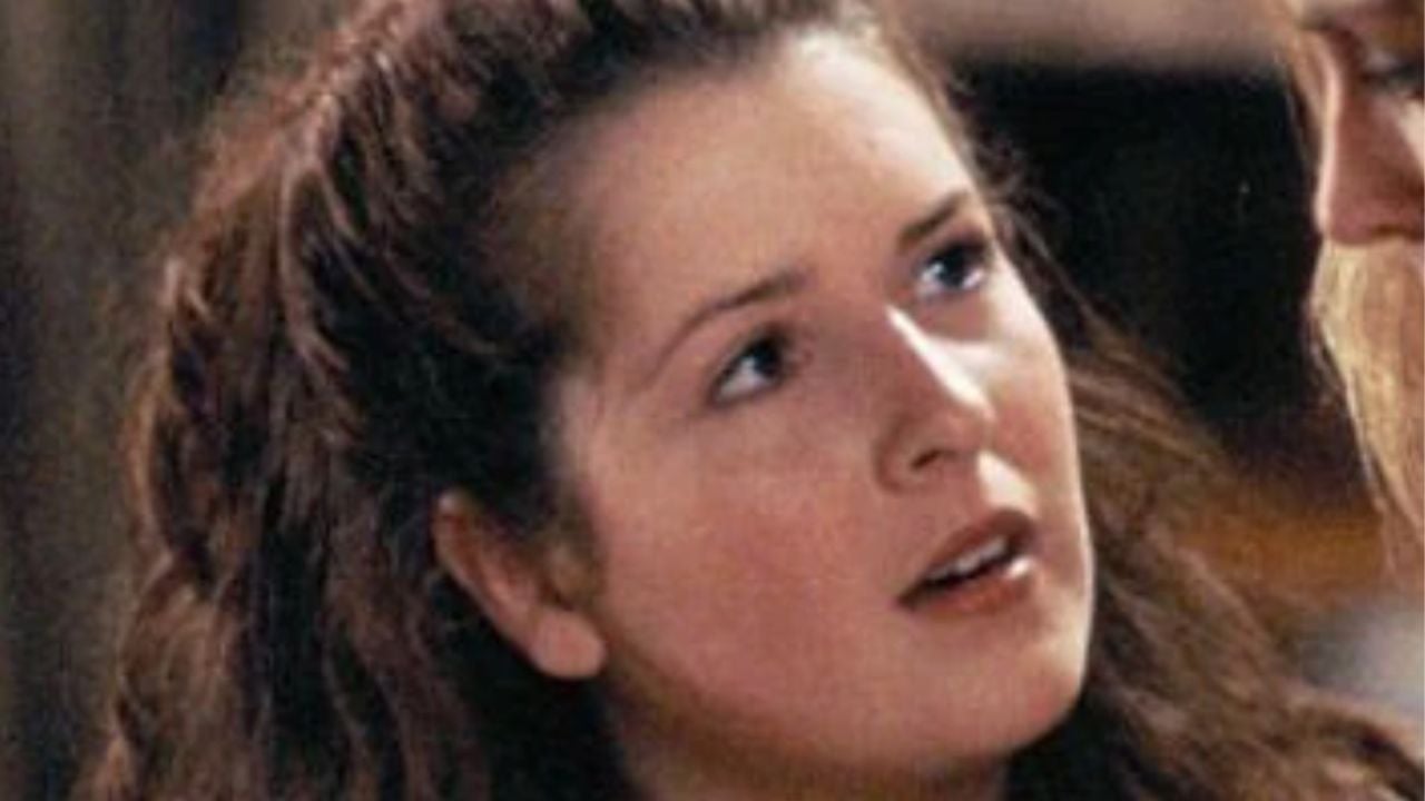 A young woman with long, wavy brown hair looks upward with a curious and concerned expression. She is indoors, and her face is softly lit.