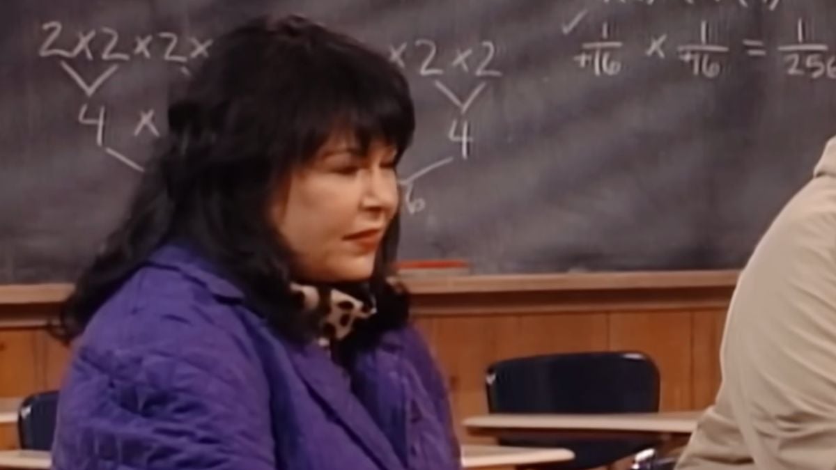 A woman with dark hair in a purple jacket sits in a classroom with a chalkboard behind her, featuring math equations written in chalk.