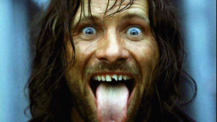 A man with long hair and a beard stares wide-eyed at the camera with his mouth open, showing missing teeth and sticking out his tongue. The background is out of focus.