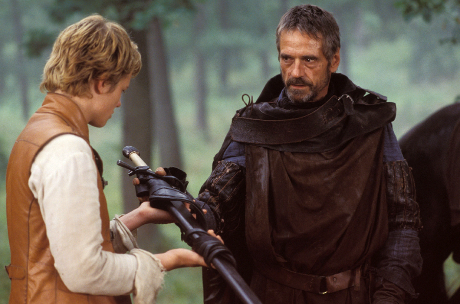 A young person in a brown vest holds a large sword, while an older man in dark medieval clothing looks at them seriously in a forest setting.
