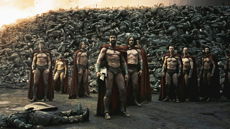 A group of muscular warriors in red capes and armor stand together in front of a massive pile of bodies, with a dramatic, cloudy sky in the background.