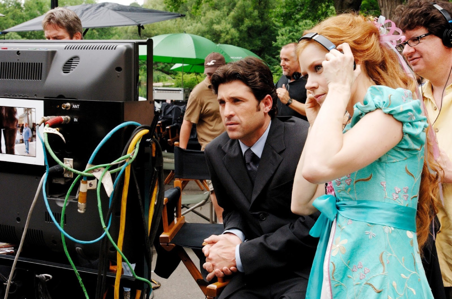 A man in a suit and a woman in a blue dress with headphones watch a monitor on a film set outdoors, surrounded by crew members and equipment.