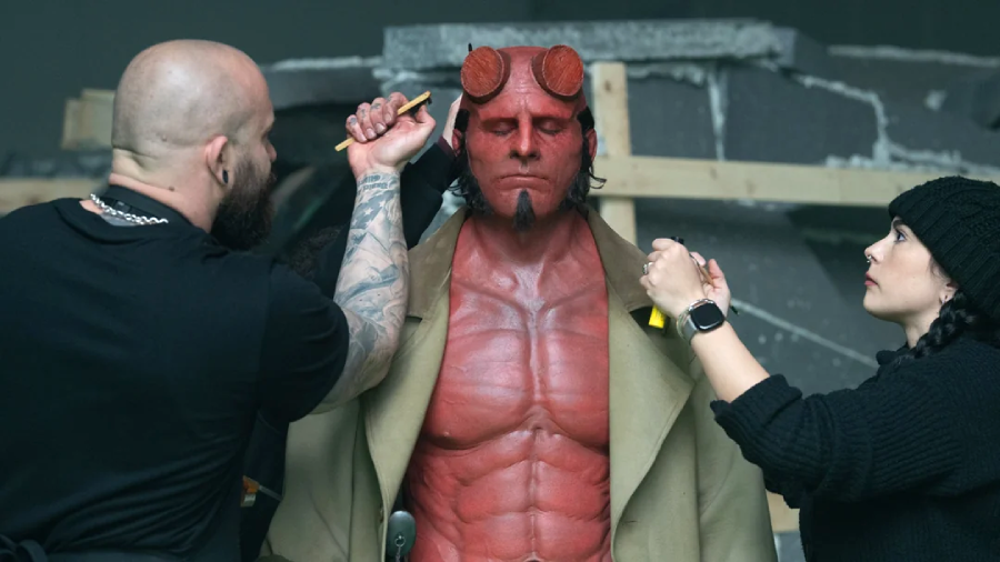 A man in detailed Hellboy makeup and costume stands still while two makeup artists touch up his face and chest on a film set with a wooden and stone-like background.