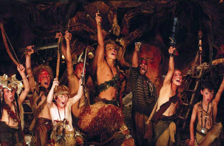 A group of children in tribal costumes cheer and raise weapons in a lively, dimly lit setting resembling a cave or hideout, with roots and natural elements in the background.