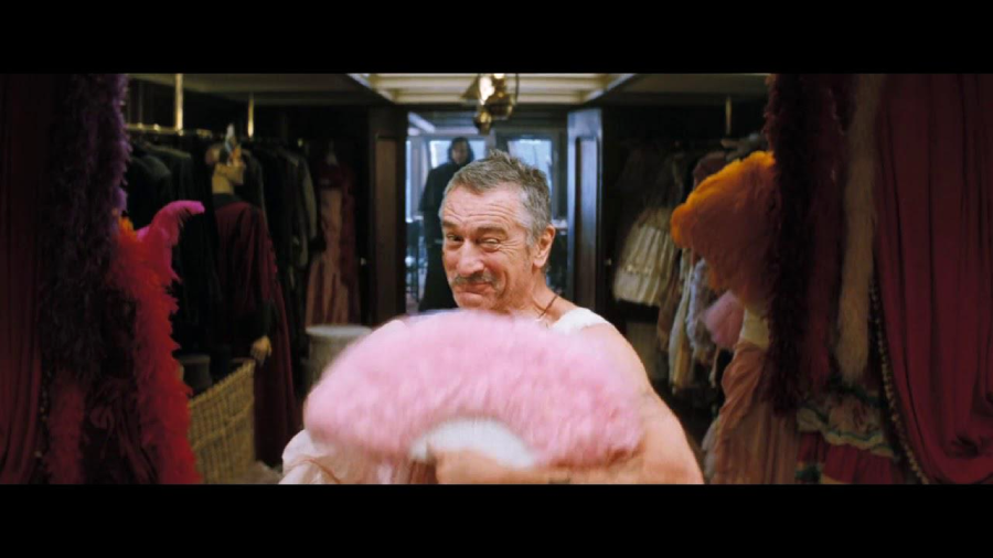 An older man stands in a dressing room holding a large pink feathered fan, smiling playfully. The room is filled with colorful costumes and clothing hanging on racks in the background.