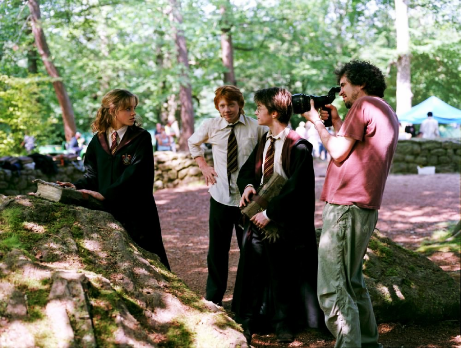 Three young actors in school uniforms stand outdoors in a wooded area, talking near a mossy rock, while a person with curly hair and a pink shirt films them with a camera. Other people and tents are visible in the background.