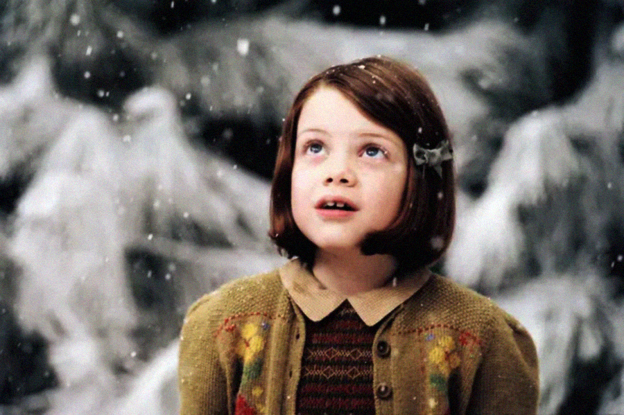 A young girl with short brown hair and a hairclip looks up in wonder as snow falls around her. She is wearing a brown sweater with colorful patterns, with a snowy, wintry background behind her.
