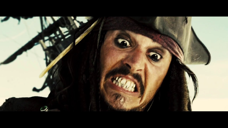 A close-up of a man dressed as a pirate with a tricorn hat, dreadlocked hair, and a beaded bandana, making a wild, exaggerated facial expression with wide eyes and bared, uneven teeth.