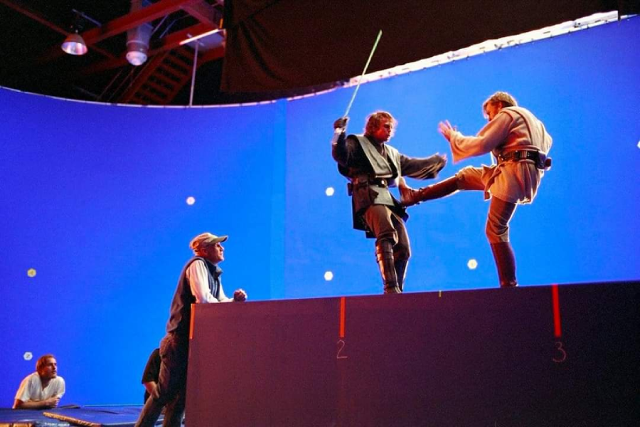 Two actors in costume perform a sword fight on a raised platform in front of a large blue screen, while crew members observe on set, likely during the filming of a movie scene with visual effects.