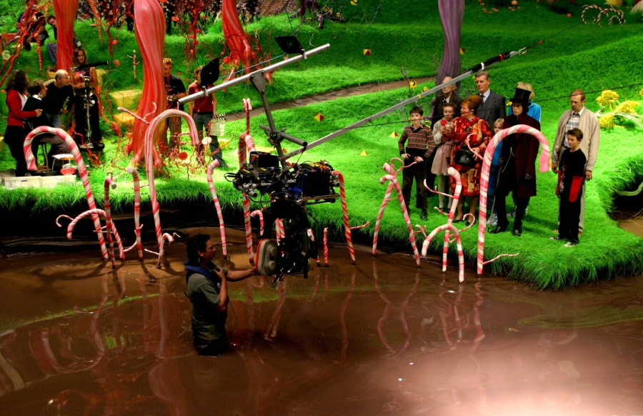 A film crew member stands in a chocolate-colored river with a camera, while actors in colorful costumes stand on candy-decorated grass in a whimsical, candy-themed set resembling Willy Wonka’s chocolate factory.