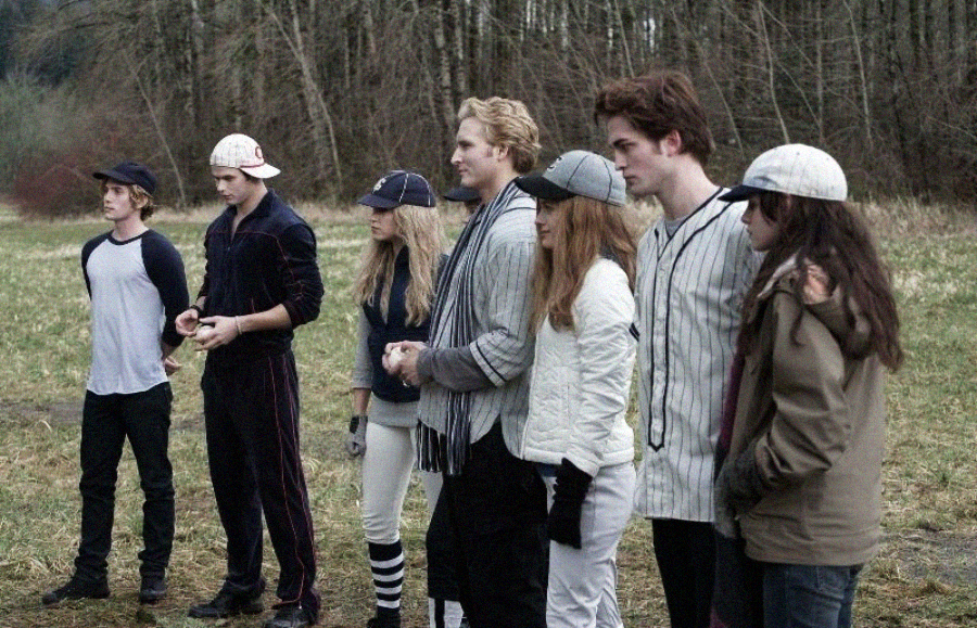 Seven people dressed in casual and baseball-themed outfits stand in a line on a grassy field, with trees in the background. Some wear hats and striped clothing, and they appear to be watching or waiting for something.