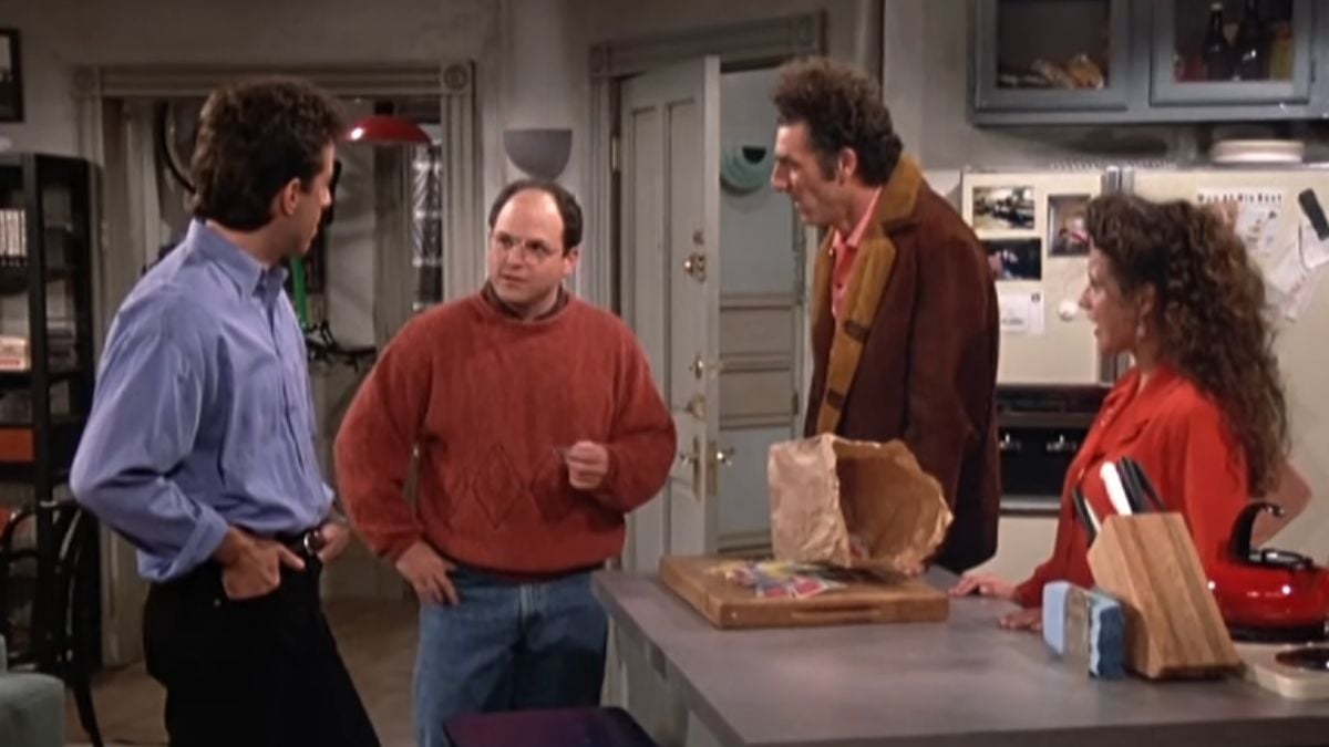 Four people are talking in a kitchen. Two stand by the counter, one wears a red sweater, and groceries and bread sit on the counter. Refrigerator and shelves are visible in the background.