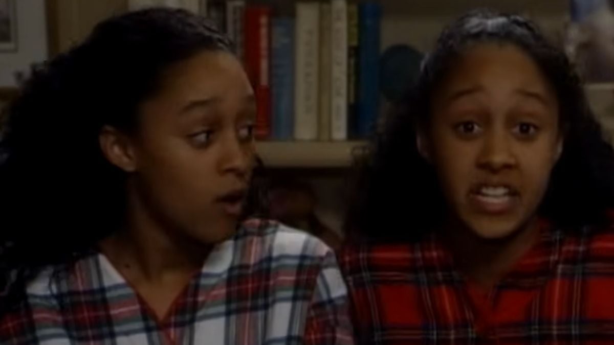 Two young women wearing matching plaid shirts sit side by side in front of a bookshelf; one looks surprised while the other is speaking.