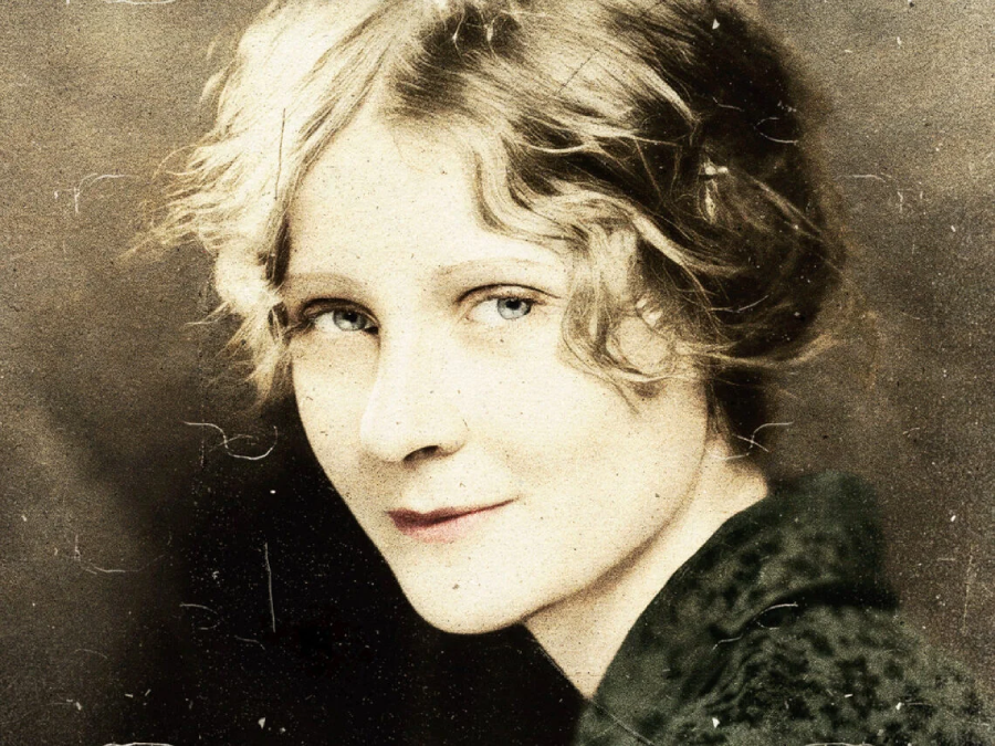 A vintage portrait of a young woman with short, wavy blonde hair and light eyes, wearing a dark patterned top, looking over her shoulder and softly smiling at the camera. The photo has a sepia tone and a slightly worn texture.