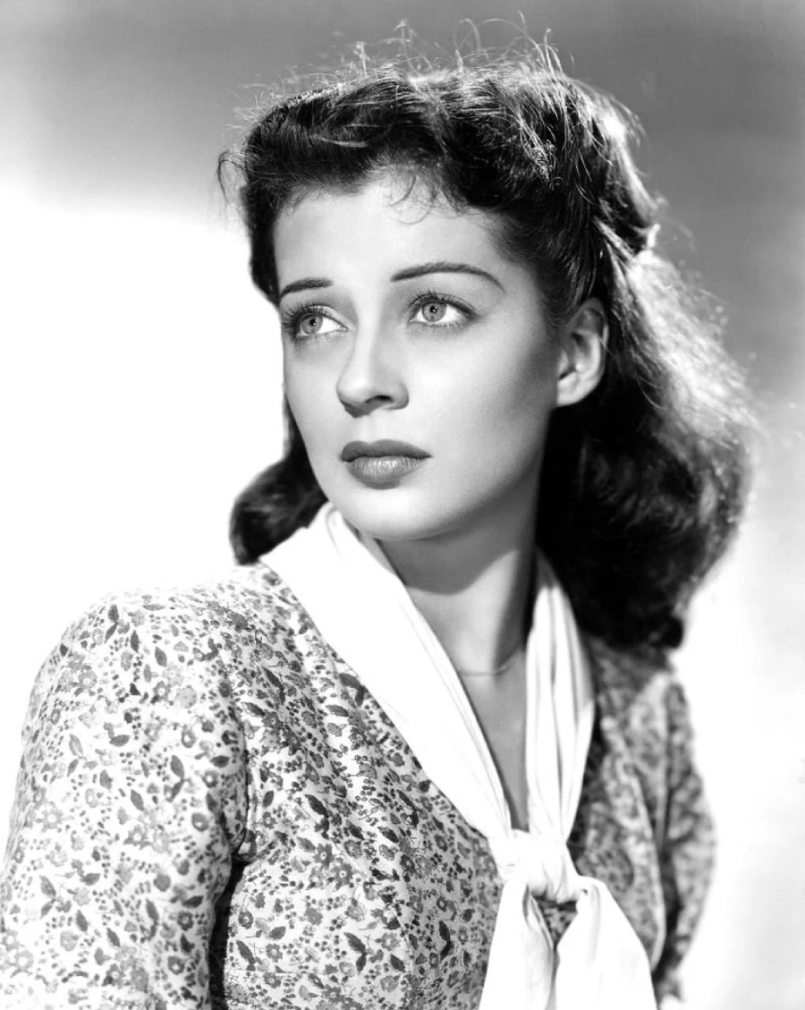 A woman with wavy dark hair and light eyes gazes slightly upward. She is wearing a patterned dress and a white scarf, with a soft, thoughtful expression in this black-and-white portrait.