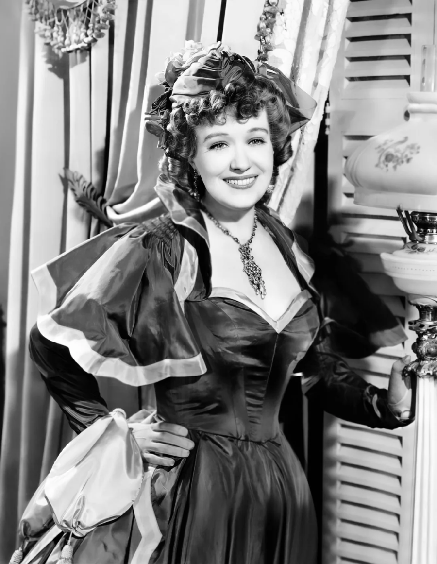 A woman in vintage, 19th-century-style clothing stands indoors, smiling. She wears a dark, off-shoulder dress with puffed sleeves, a large necklace, and a decorative hat, with her hand resting on a lamp near window blinds.