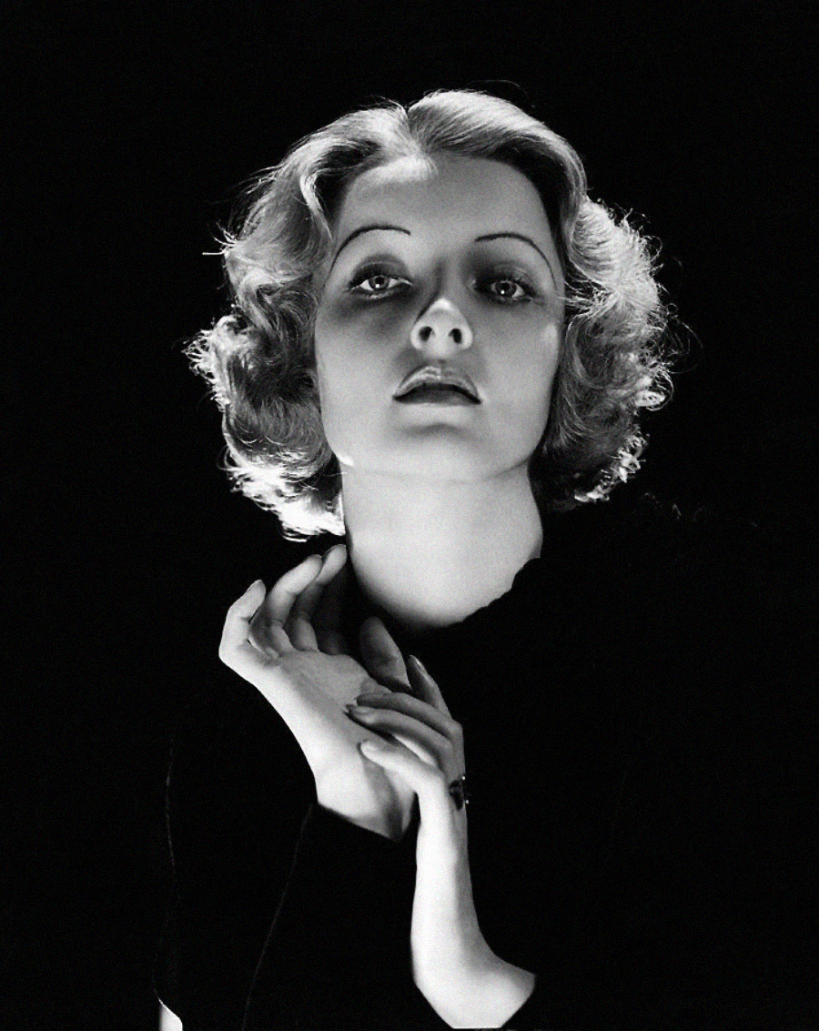 A woman with short, wavy blonde hair poses dramatically against a dark background, gazing slightly upward with her hands gracefully positioned near her face and neck, illuminated by soft, high-contrast lighting.