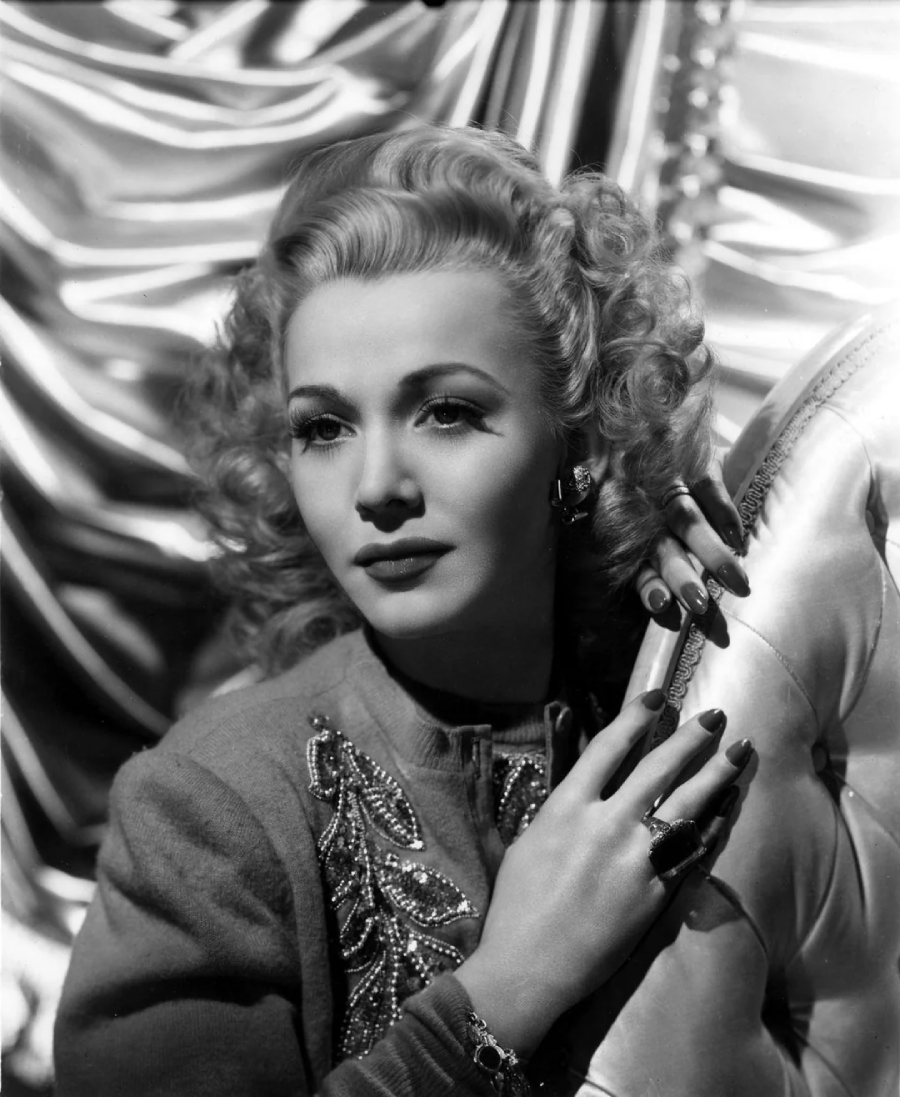 Black-and-white portrait of a woman with wavy blonde hair, wearing a detailed top with embroidery, long nails, and rings, resting her hand on a shiny, tufted chair with a satin backdrop behind her.