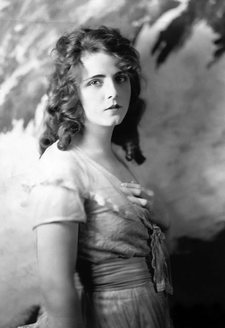 A young woman with curly hair in a light, vintage dress stands with one hand on her chest, looking softly at the camera against a blurred, artistic backdrop.