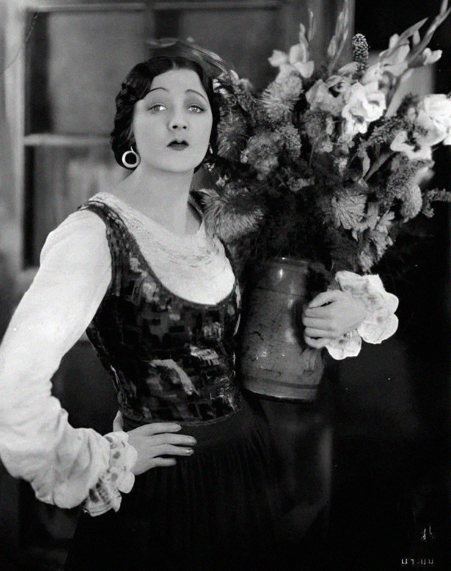 A woman with short dark hair and hoop earrings holds a large vase of flowers. She wears a long-sleeved blouse with a velvet vest and skirt, gazing slightly upward with a calm expression. The background is softly blurred.