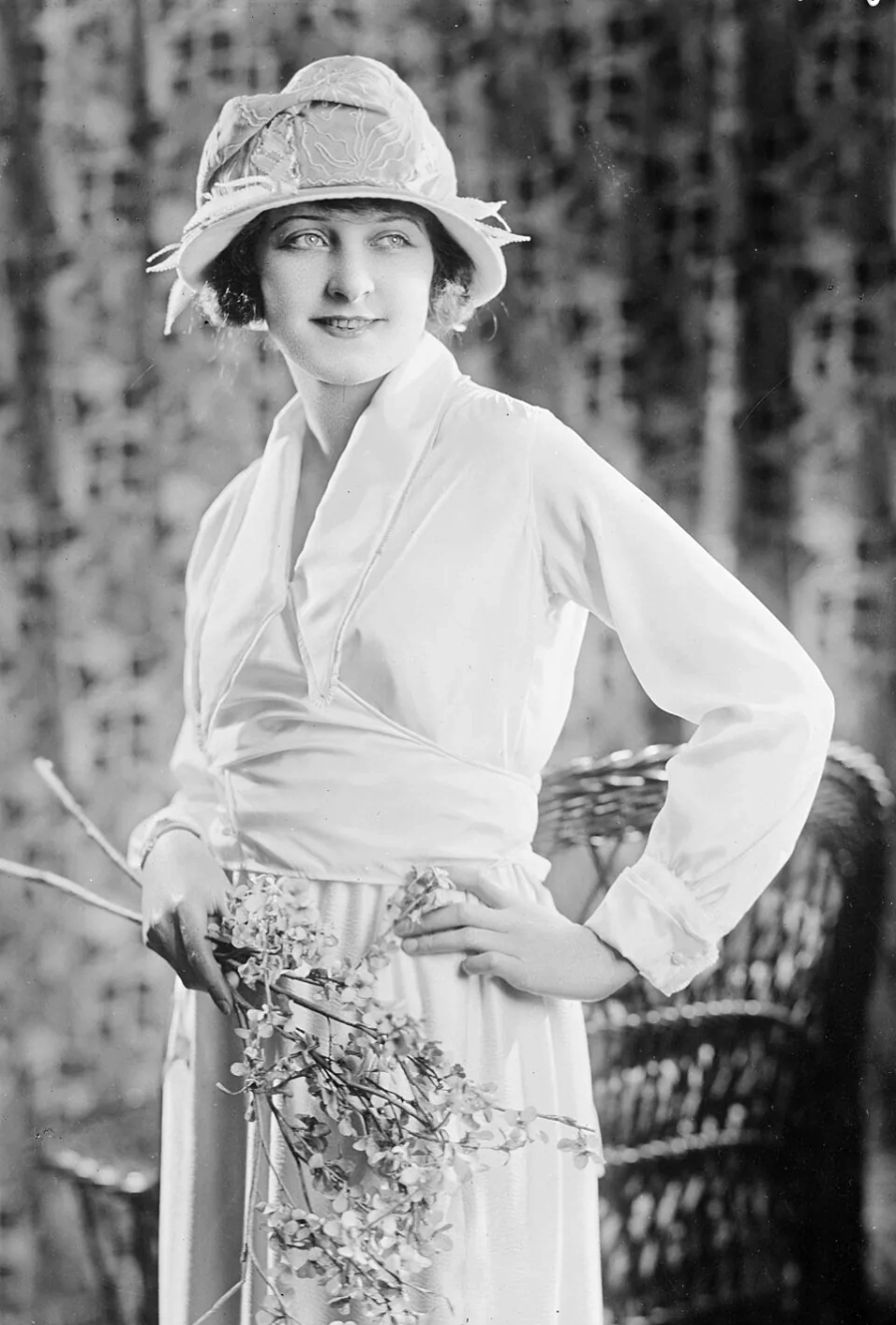 A woman in a long-sleeved white dress and wide-brimmed hat stands holding a branch with small flowers, gazing to the side and smiling, with a wicker chair and patterned curtain in the background.