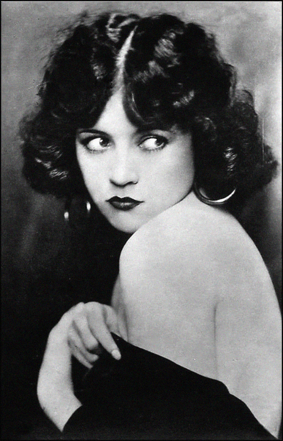 Black and white portrait of a woman with wavy dark hair, large hoop earrings, and dark lipstick. She glances to her right over her bare shoulder, holding a garment against her chest.