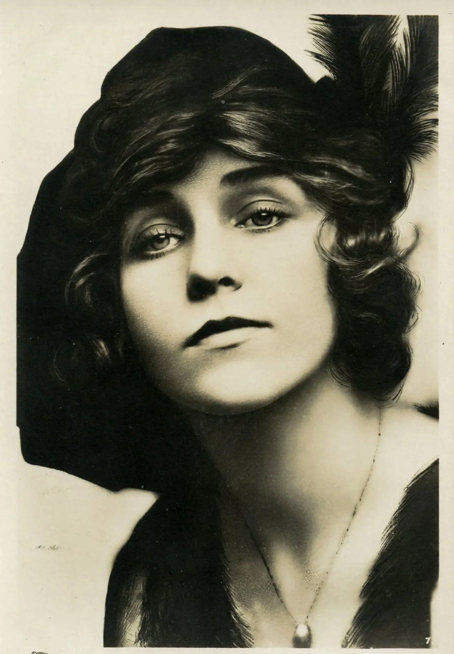 Vintage black-and-white portrait of a woman with wavy hair, wearing a dark hat adorned with a feather, a necklace, and a dark outfit, gazing slightly upward with a neutral expression.