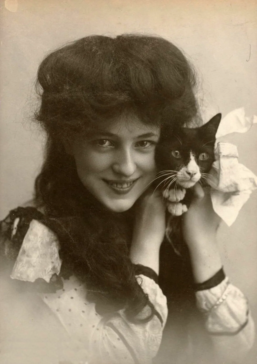 A vintage black-and-white photo of a smiling young woman with voluminous hair holding a black-and-white cat close to her face. The cat has a ribbon tied around its neck.
