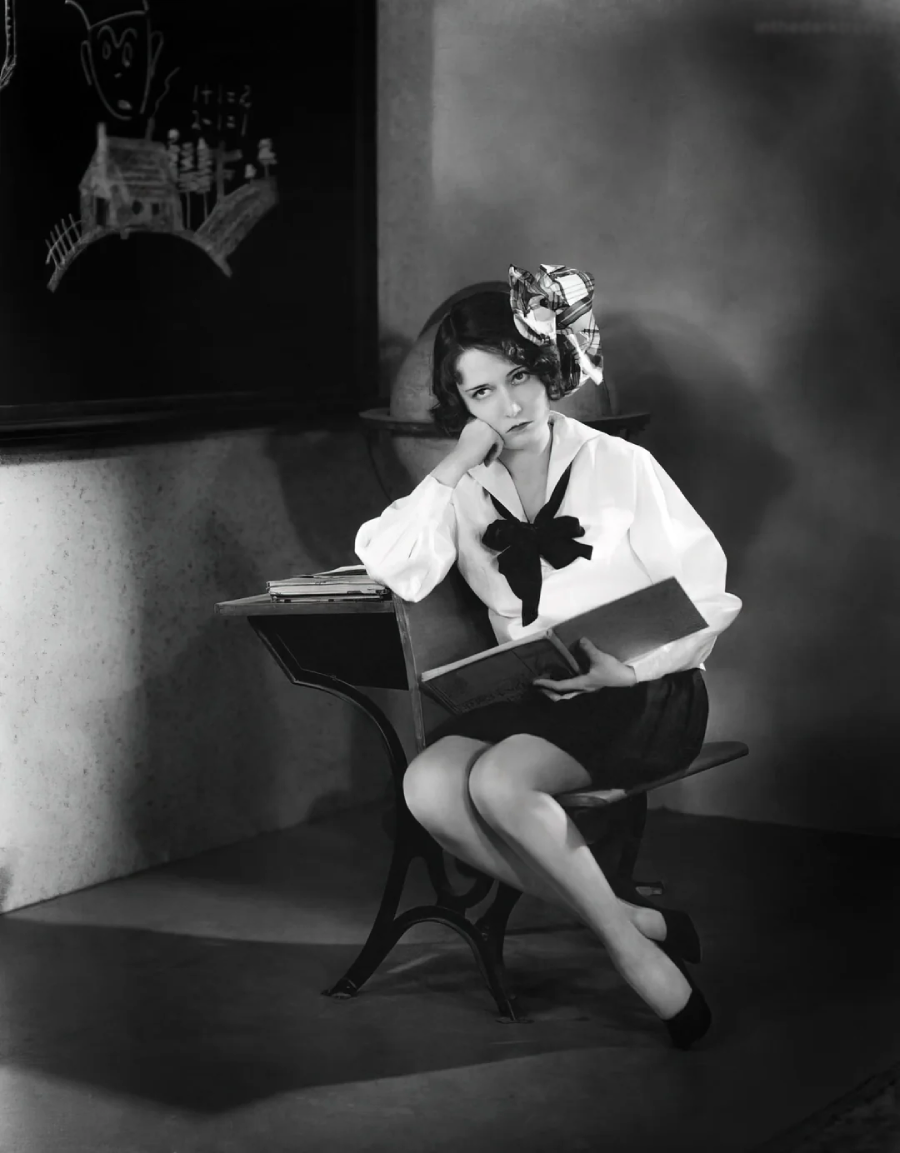 A young woman in vintage clothing sits at an old-fashioned school desk, resting her face on her hand and looking bored. She holds an open book and wears a large bow in her hair. A chalkboard with drawings is behind her.