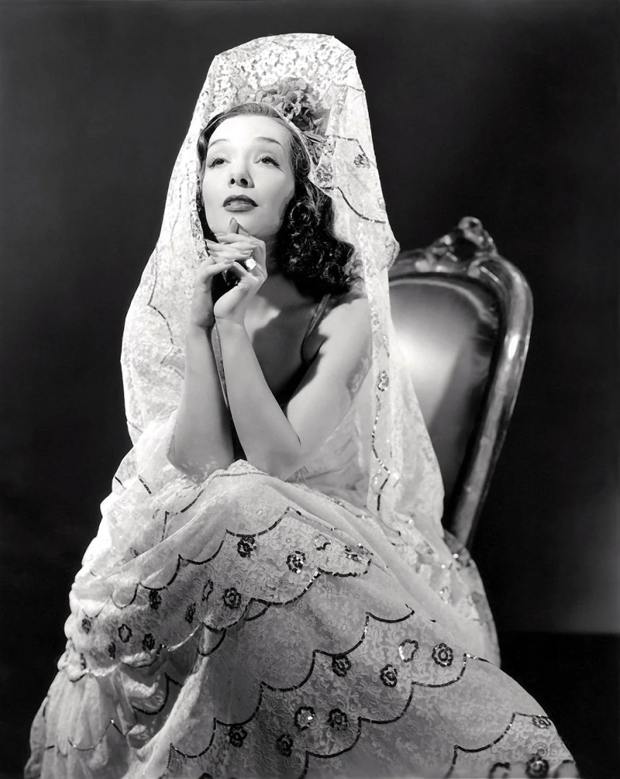 A woman in a vintage lace gown and veil sits on an ornate chair, hands clasped under her chin, gazing upward. The black-and-white photo conveys a classic, glamorous, old Hollywood style.