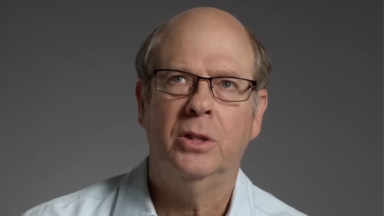 A middle-aged man with glasses, a receding hairline, and wearing a light blue shirt looks slightly upward against a plain gray background.