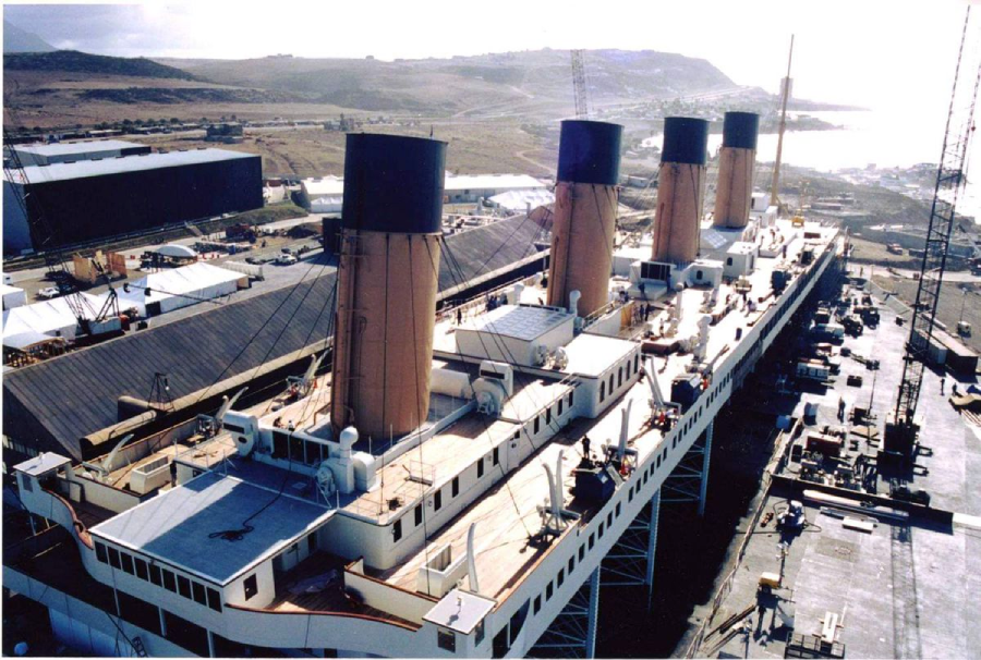 A large ocean liner with four smokestacks is docked at a shipyard, surrounded by cranes and industrial buildings, with hills and coastline visible in the background.