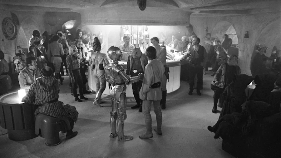 A busy cantina filled with various aliens, humans, and two robots, C-3PO and R2-D2, in a lively, dimly lit space with curved walls and a central bar.