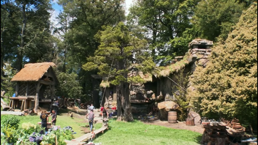 A fantasy-style village set among tall trees, featuring thatched-roof cottages, stone structures, and people walking along wooden paths surrounded by greenery and garden beds.