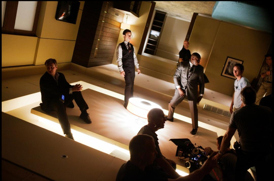 Several actors in suits stand or sit on a brightly lit, modern set, while crew members with equipment work in the foreground; the set appears tilted, creating a surreal, disorienting effect.