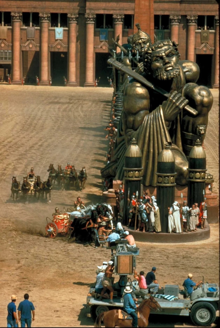 A movie set features a chariot race with horses and riders circling giant warrior statues. Crew members, cameras, and equipment are visible in the foreground, while actors in costume and grand columns appear in the background.