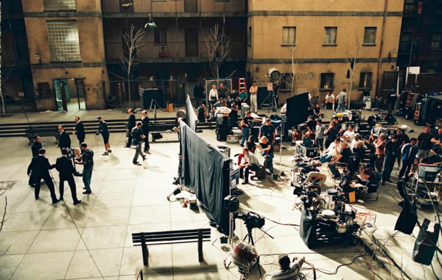 A busy film set in an outdoor courtyard, with crew members, actors, lighting equipment, cameras, and various production gear scattered around as people prepare for a shoot. The surrounding buildings are visible in the background.