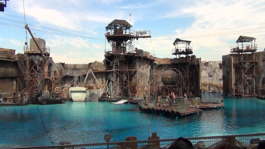 A large outdoor water stage set with rusted metal structures, towers, and platforms surrounds a blue pool, designed to resemble an industrial post-apocalyptic scene. Performers stand on floating platforms.