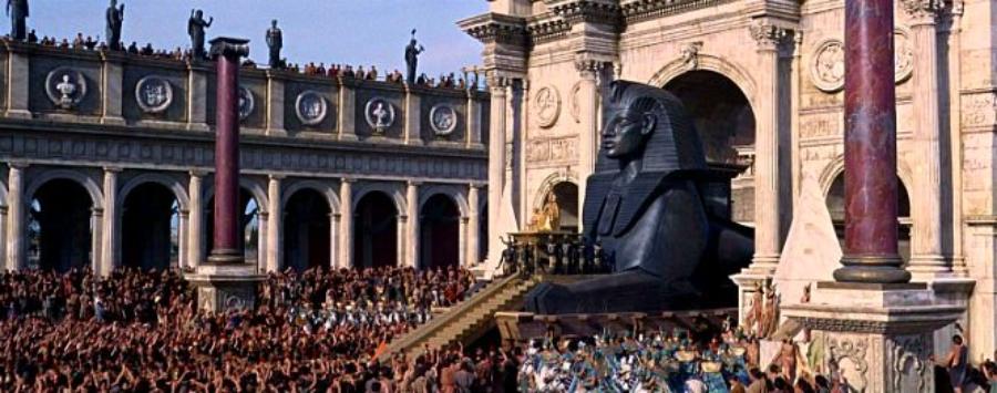 A grand ancient scene with crowds gathered before a large stone sphinx statue, grand columns, and an ornate marble archway, evoking a sense of historical or cinematic spectacle.