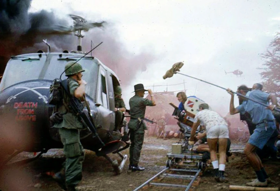 A film crew records a scene with actors dressed as soldiers near a military helicopter marked "Death From Above," surrounded by smoke and equipment on a track.