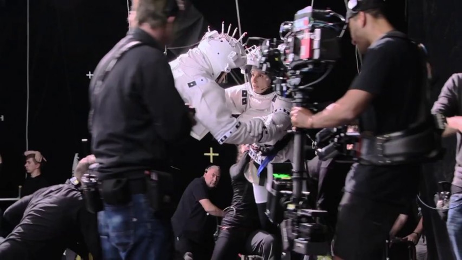 Behind-the-scenes view of a film set, showing crew members operating cameras while an actor in a white astronaut suit performs a scene. Several people in black clothing work around the set with equipment.
