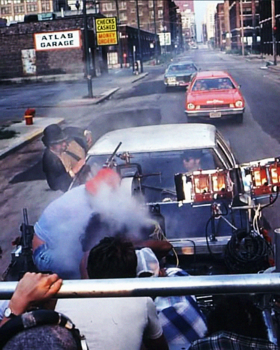 A film crew shoots a scene with cameras and lights mounted on a car in an urban street. Smoke rises from the vehicle as a red car follows behind. City buildings and signs are visible in the background.