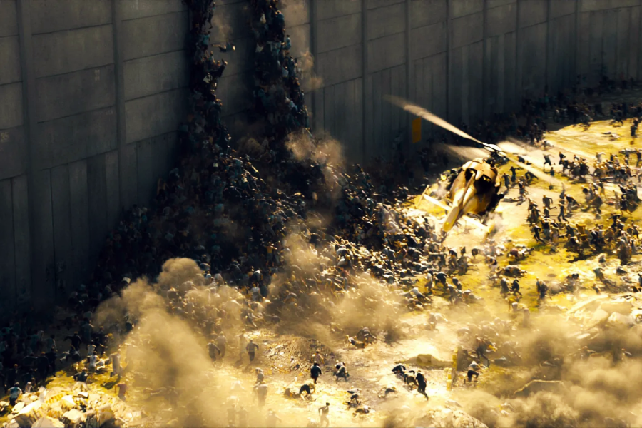 A helicopter flies over a chaotic scene as a massive crowd climbs on top of each other, scaling a tall concrete wall, with dust and debris filling the air.
