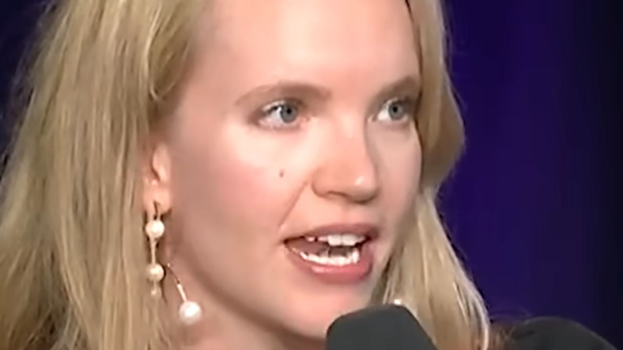 A woman with long blonde hair speaks into a microphone. She is wearing dangling pearl earrings and is shown against a dark background.