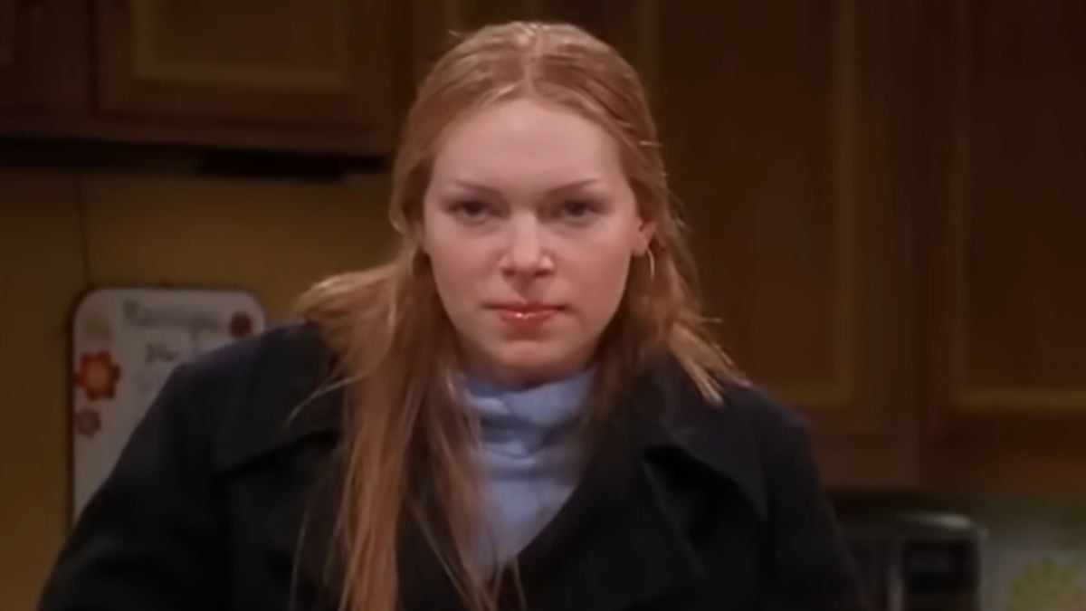 A woman with long, straight red hair and a serious expression sits indoors, wearing a black coat and light blue turtleneck, with a kitchen background behind her.