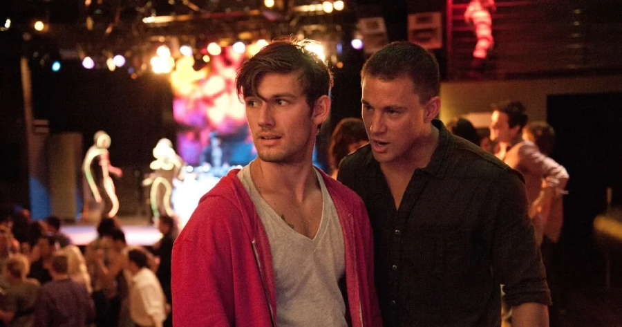 Two men stand close together in a crowded nightclub. One wears a red hoodie and looks ahead, while the other, in a dark shirt, leans in and speaks to him. Colorful stage lights illuminate the background with people dancing.