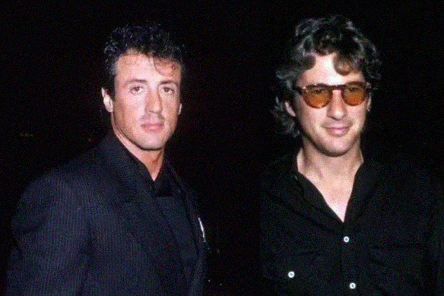 Two men stand side by side against a dark background, both wearing black outfits. One has short dark hair and a serious expression, while the other has wavy hair, glasses, and a slight smile.