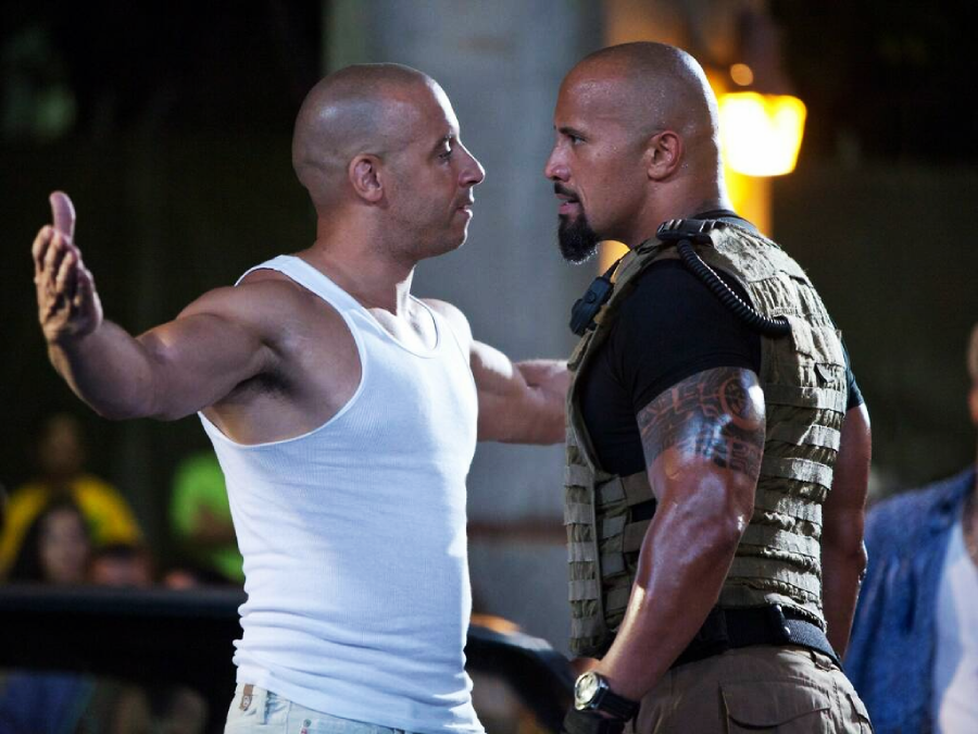 Two muscular men stand face-to-face in an intense confrontation; one wears a white tank top with arms outstretched, the other wears a tactical vest. Both appear serious, with a blurred crowd in the background.