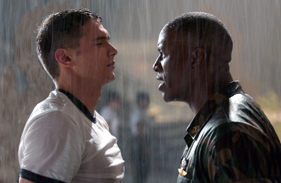 Two men stand face to face in the rain, one in a wet white T-shirt, the other in a dark uniform, appearing to have an intense confrontation.